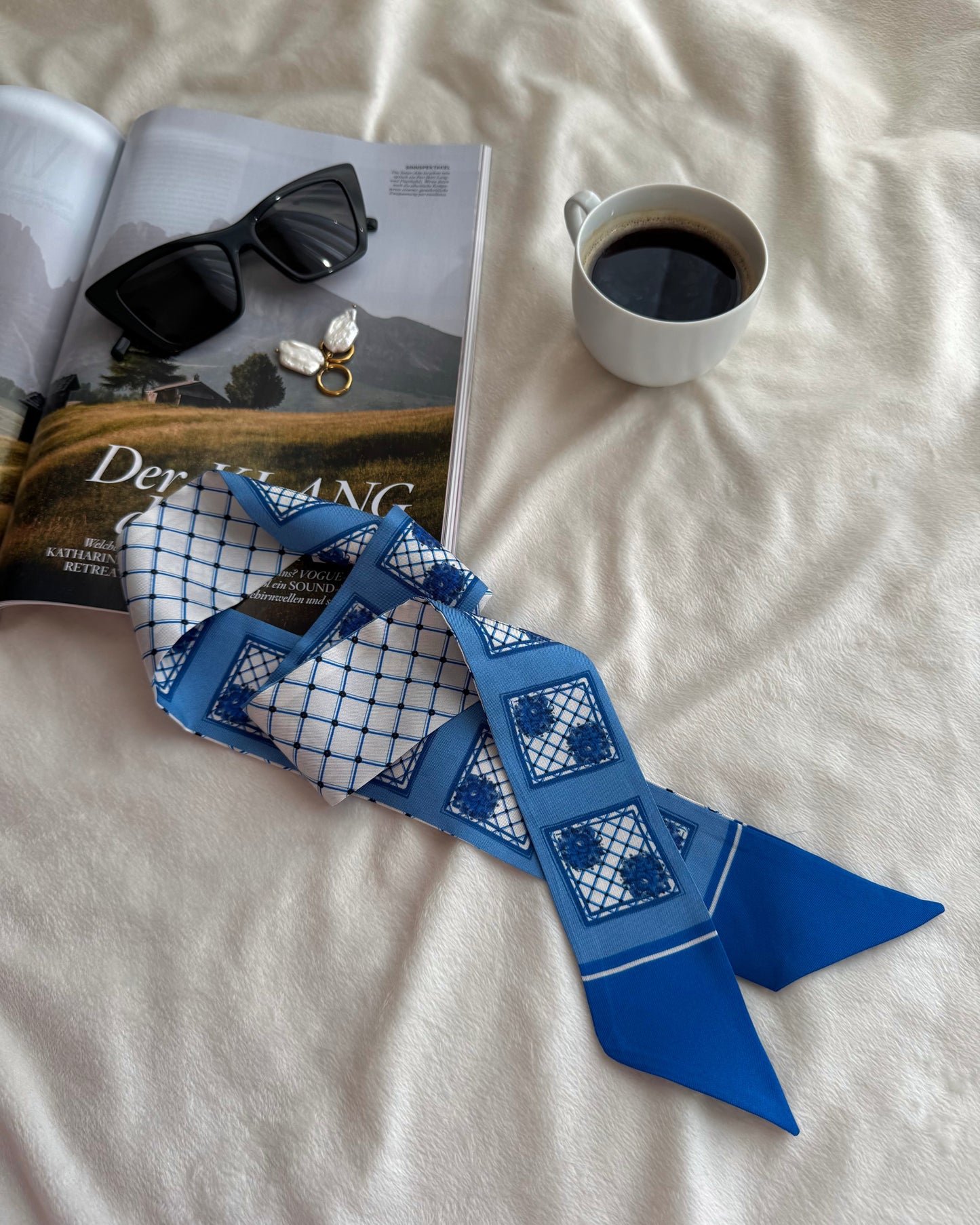 Blue patterned scarf on a white surface with a magazine, sunglasses, and a cup of coffee.